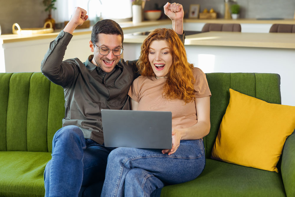 Excited man and woman sitting on a couch with a laptop in their lap holding their arms up in victory for their loan approval